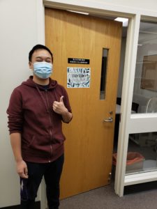 Student standing in front of Study Room 1 with a thumbs up sign. The Study Room has a wooden door and glass windows to the right of the door.